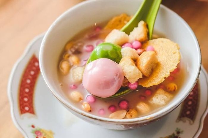 Gambar Es Sekoteng yang disajikan dalam mangkuk besar, penuh dengan potongan buah-buahan segar, kacang hijau, dan potongan roti, semua disiram dengan sirup jahe yang kental dan berwarna coklat muda